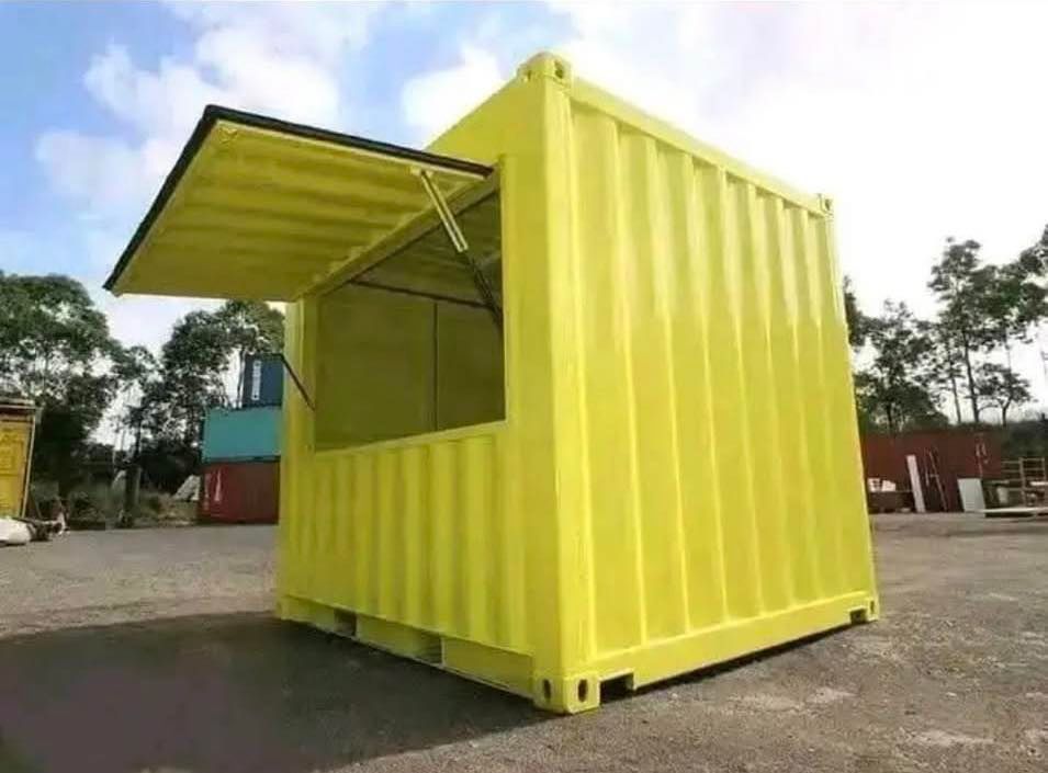 A bright yellow shipping container converted into a small kiosk with a large, hinged service window open on the side.