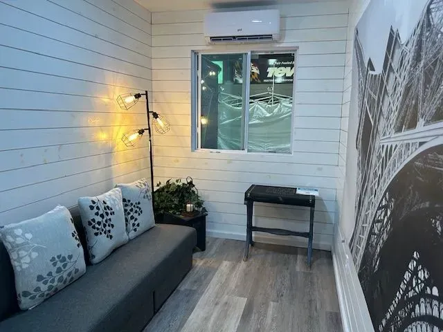 A cozy small room with white shiplap walls, a gray sofa, a floor lamp, a window, a mini-split AC, and wall art.