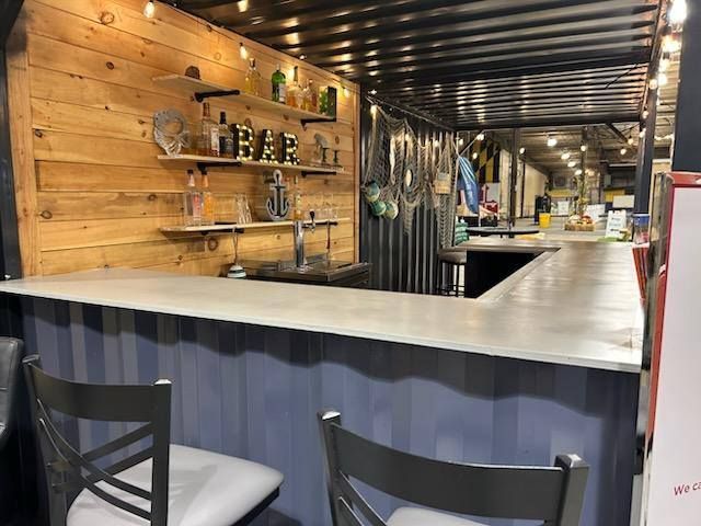 A rustic bar built inside a shipping container featuring a wood-paneled wall, open shelving, and two gray bar stools.