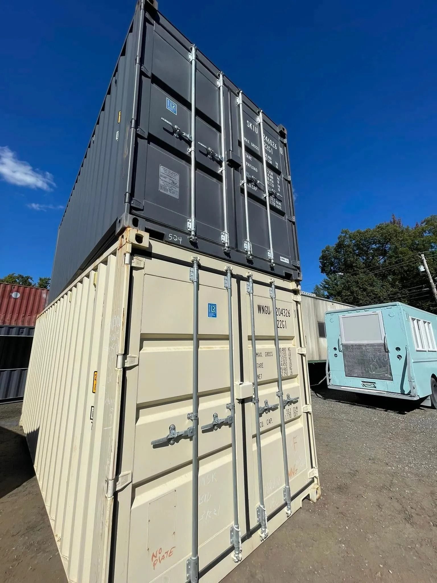 A dark gray shipping container stacked on top of a tan shipping container outdoors against a clear blue sky.