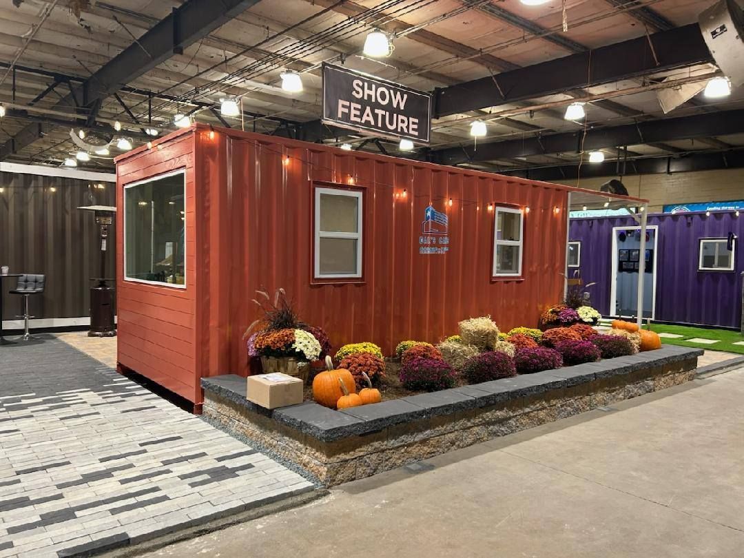 An orange repurposed shipping container used as a show exhibit, decorated with autumn flowers and pumpkins on a stone base.