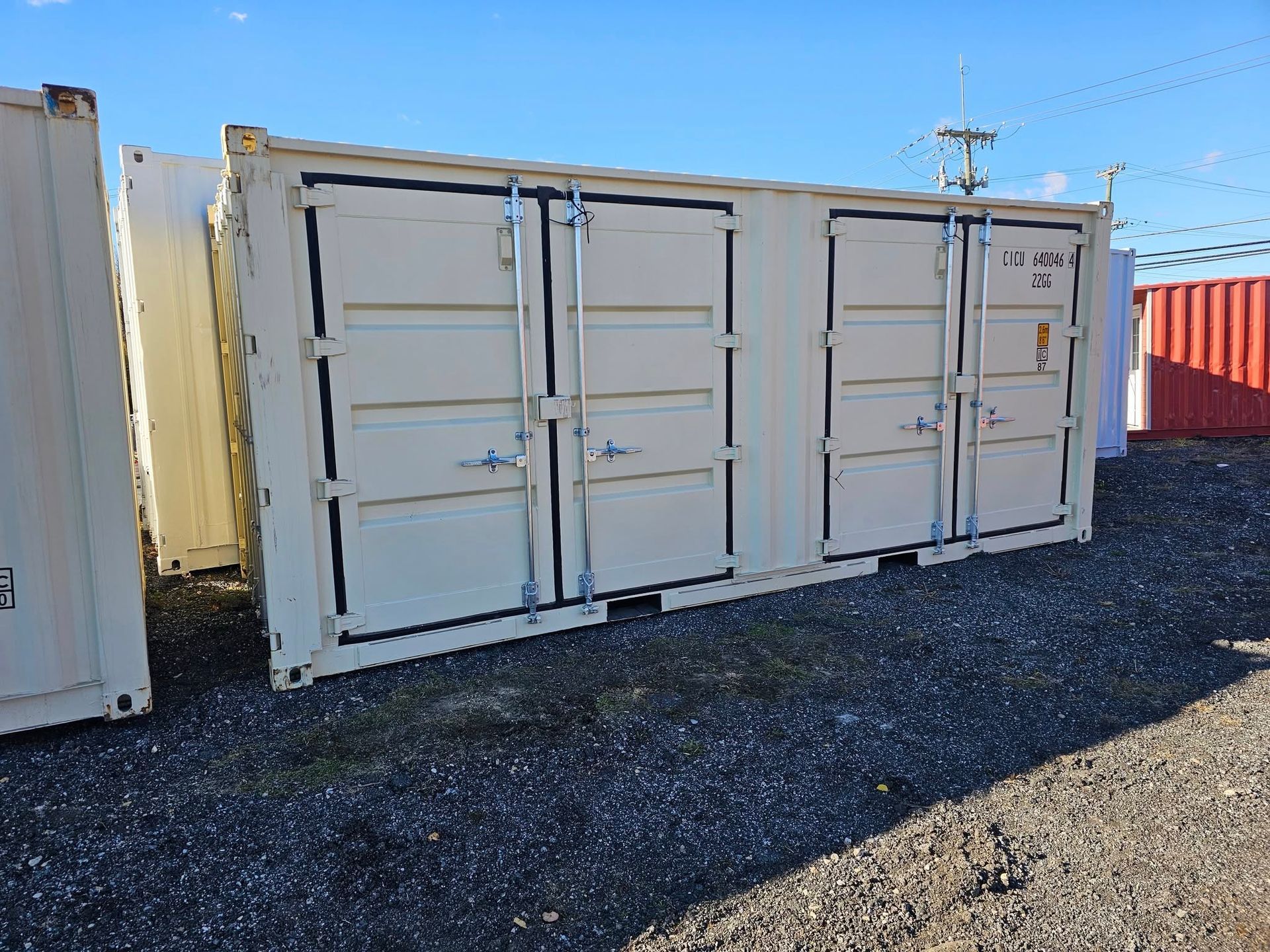 Beige shipping containers with double doors arranged in rows on a gravel lot under a clear blue sky.