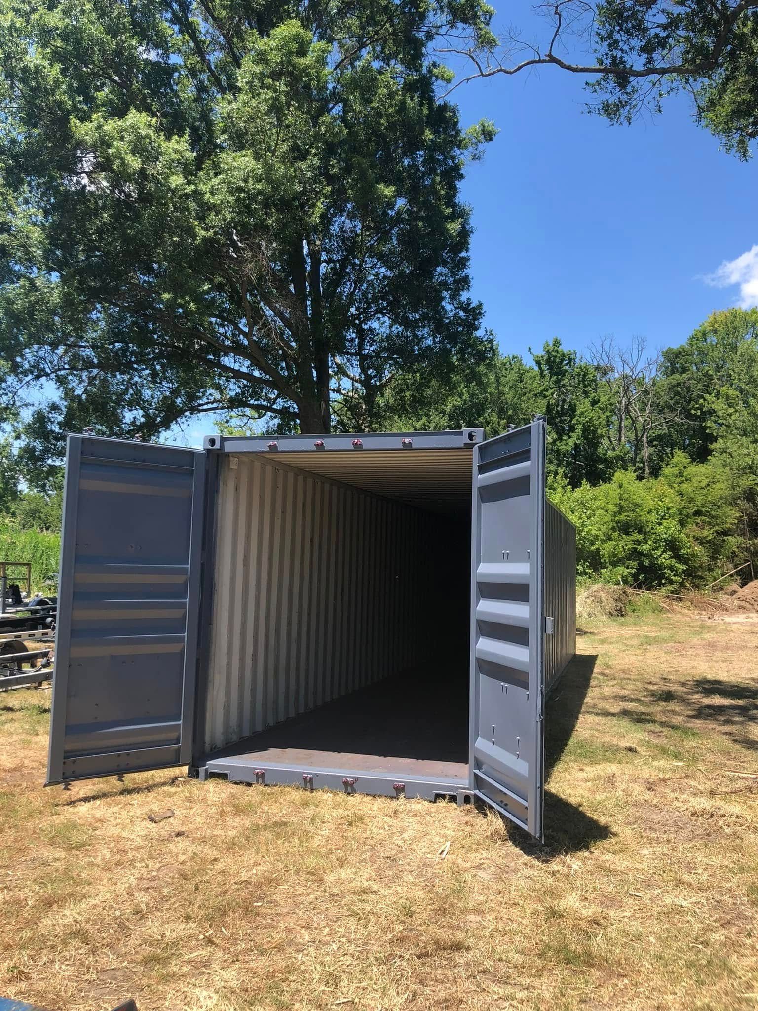 A grey shipping container with open doors sits in a grassy, wooded area under a blue sky.