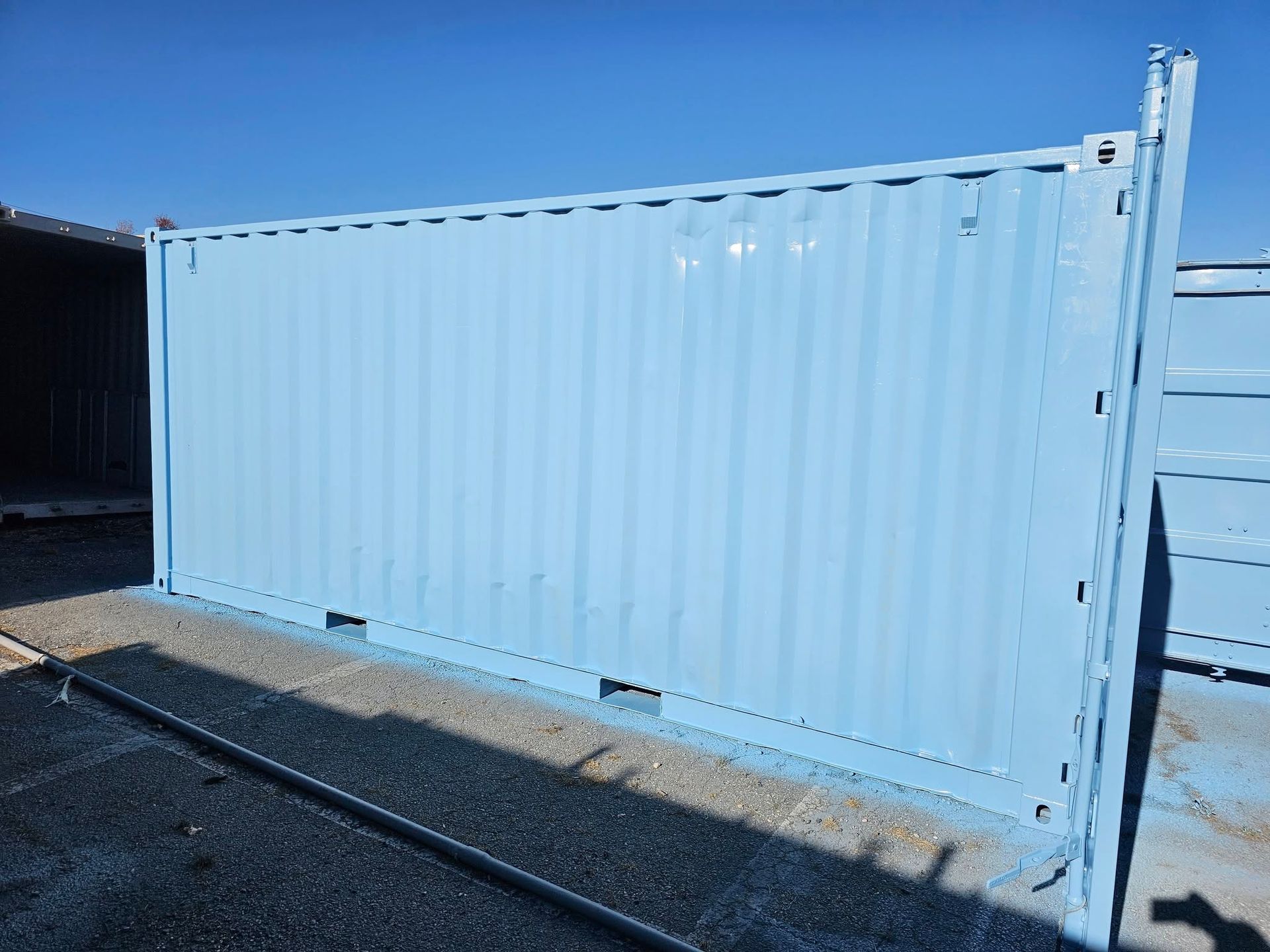 A light blue shipping container sits on a gravel lot under a clear blue sky.