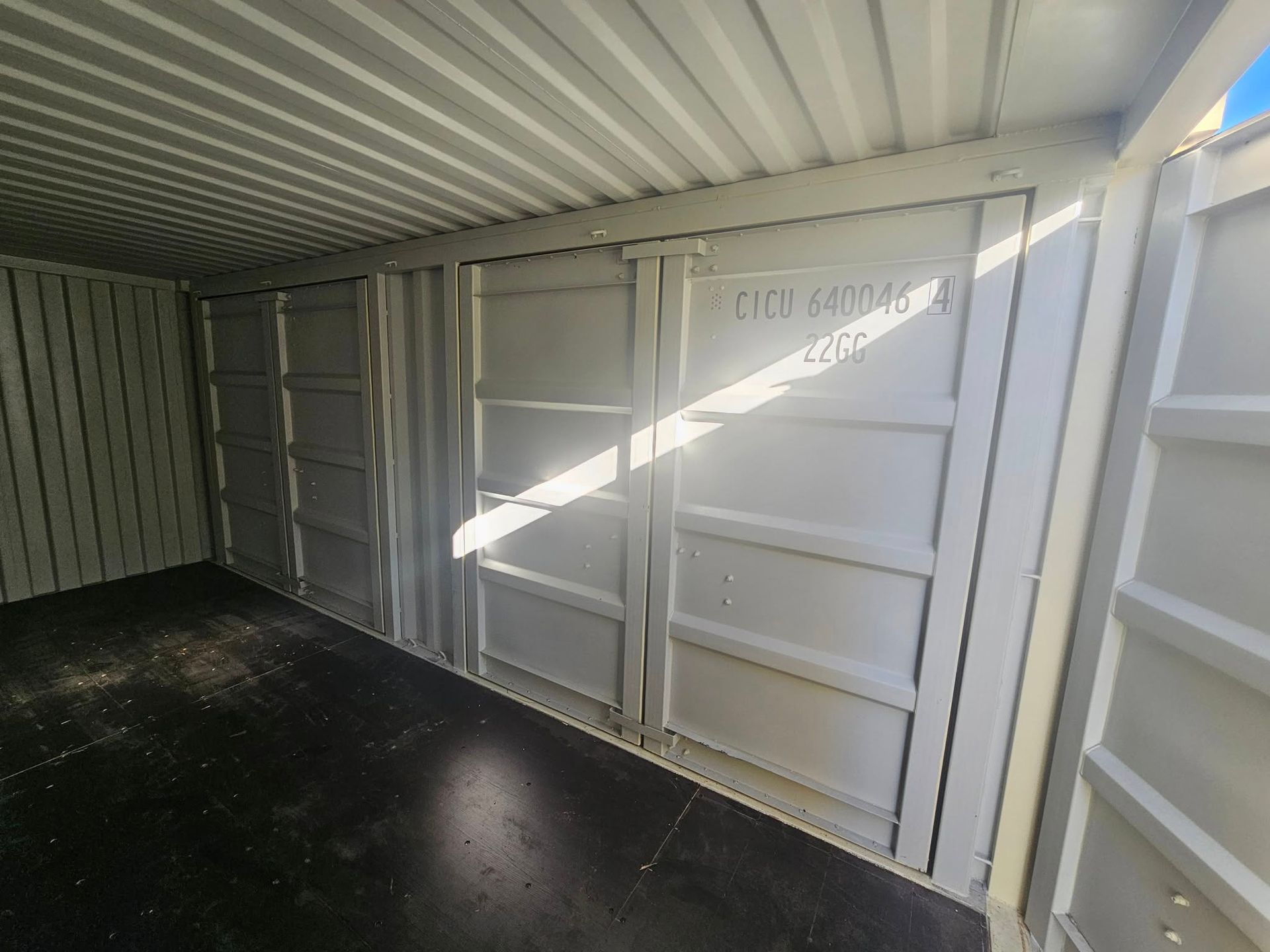 Inside a shipping container with grey metal walls, a dark floor, and sunlight streaming in through the open doorway.
