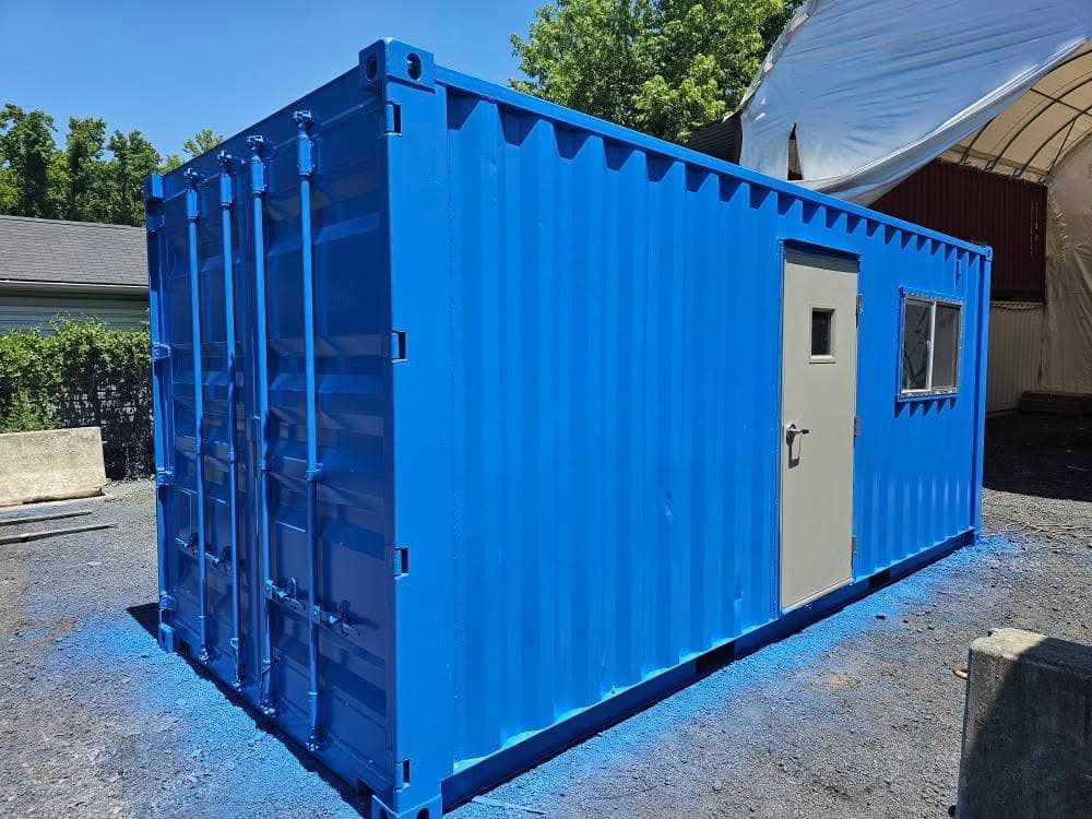A bright blue modified shipping container with a single door and window, sitting on a gravel lot under a clear blue sky.