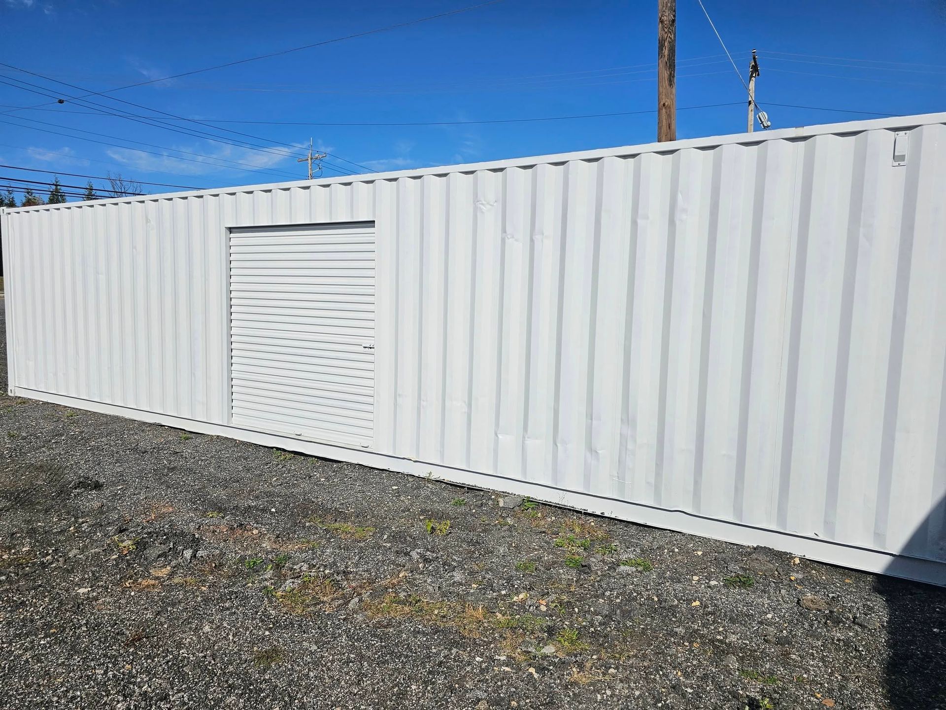 A white shipping container with a closed roll-up metal door, positioned on a gravel surface under a clear blue sky.