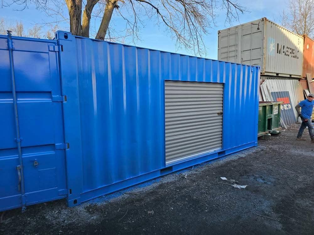 A bright blue shipping container with a silver roll-up door, parked on a gravel lot near other containers and a tree.