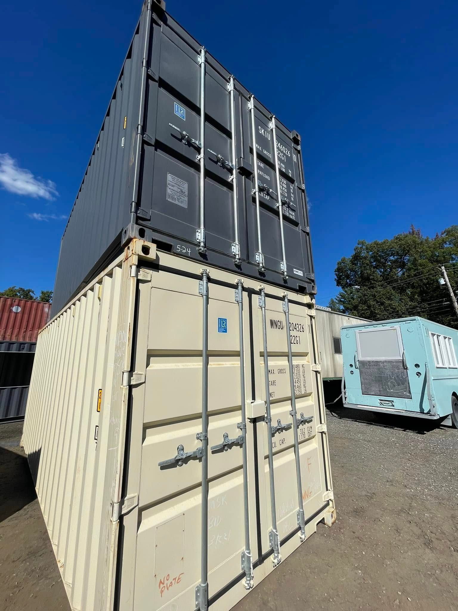 A tan shipping container stands on the ground with a dark gray container stacked on top, set against a clear blue sky.