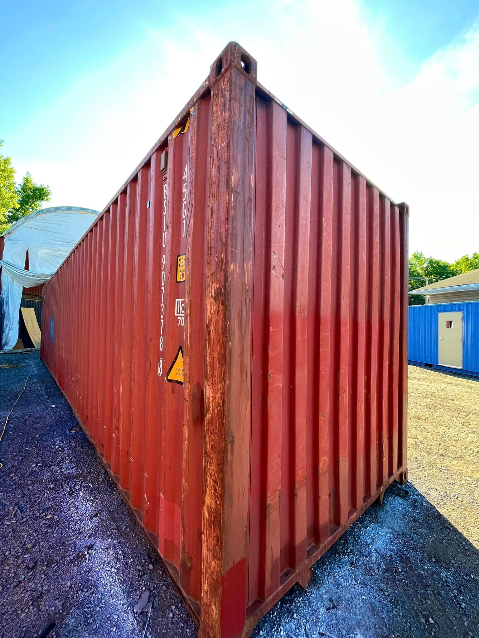 A rusted red shipping container stands on a gravel lot under a bright, sunny sky.