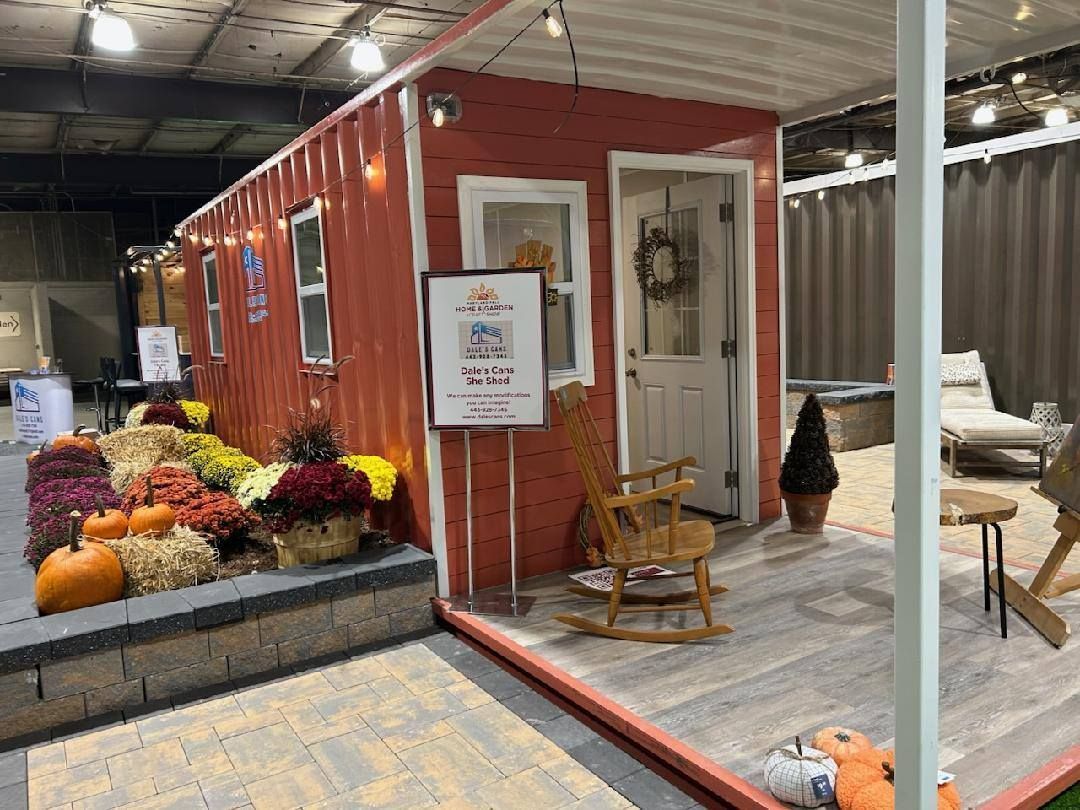 A red, barn-style tiny home display with flowers, pumpkins, a rocking chair, and a white front door on a porch.