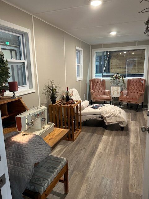 A light-filled room features a sewing machine on a wooden desk, a cushioned chaise lounge, and two velvet pink armchairs.