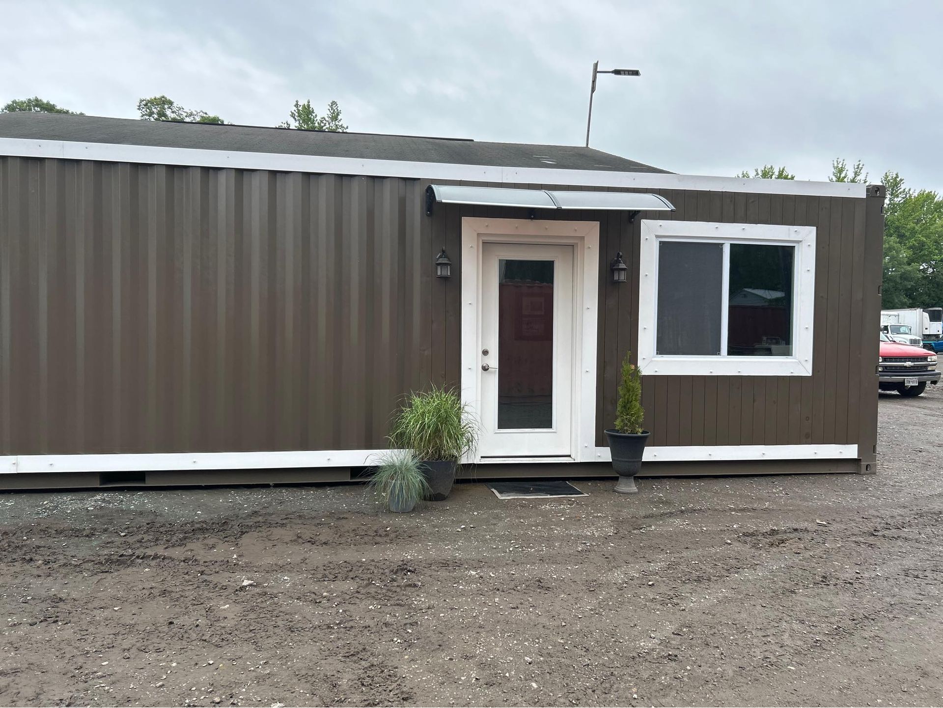 A brown shipping container building on a gravel lot, featuring a single white-trimmed door and a matching window.