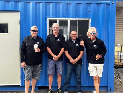 Four people wearing matching black shirts stand in front of a blue shipping container building.