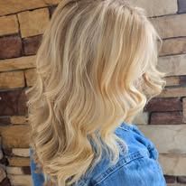 A woman with blonde hair and a denim jacket is standing in front of a brick wall.