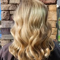 A woman with long blonde wavy hair is standing in front of a brick wall.