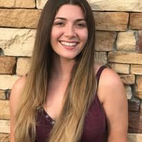 A woman with long hair is smiling in front of a brick wall.