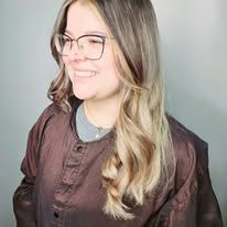 A woman wearing glasses and a brown jacket is smiling.