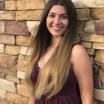A woman with long hair is standing in front of a brick wall and smiling.