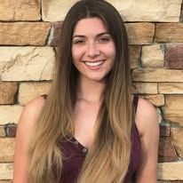 A woman with long hair is smiling in front of a brick wall.