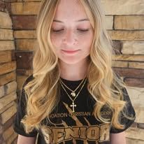 A woman with long blonde hair is wearing a black shirt and a necklace.