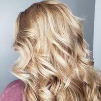 A woman with long blonde wavy hair is wearing a pink shirt.