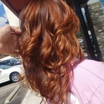 A woman with long red hair is standing in front of a car in a parking lot.