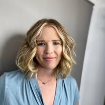 A woman with blonde hair is wearing a blue shirt and a necklace.