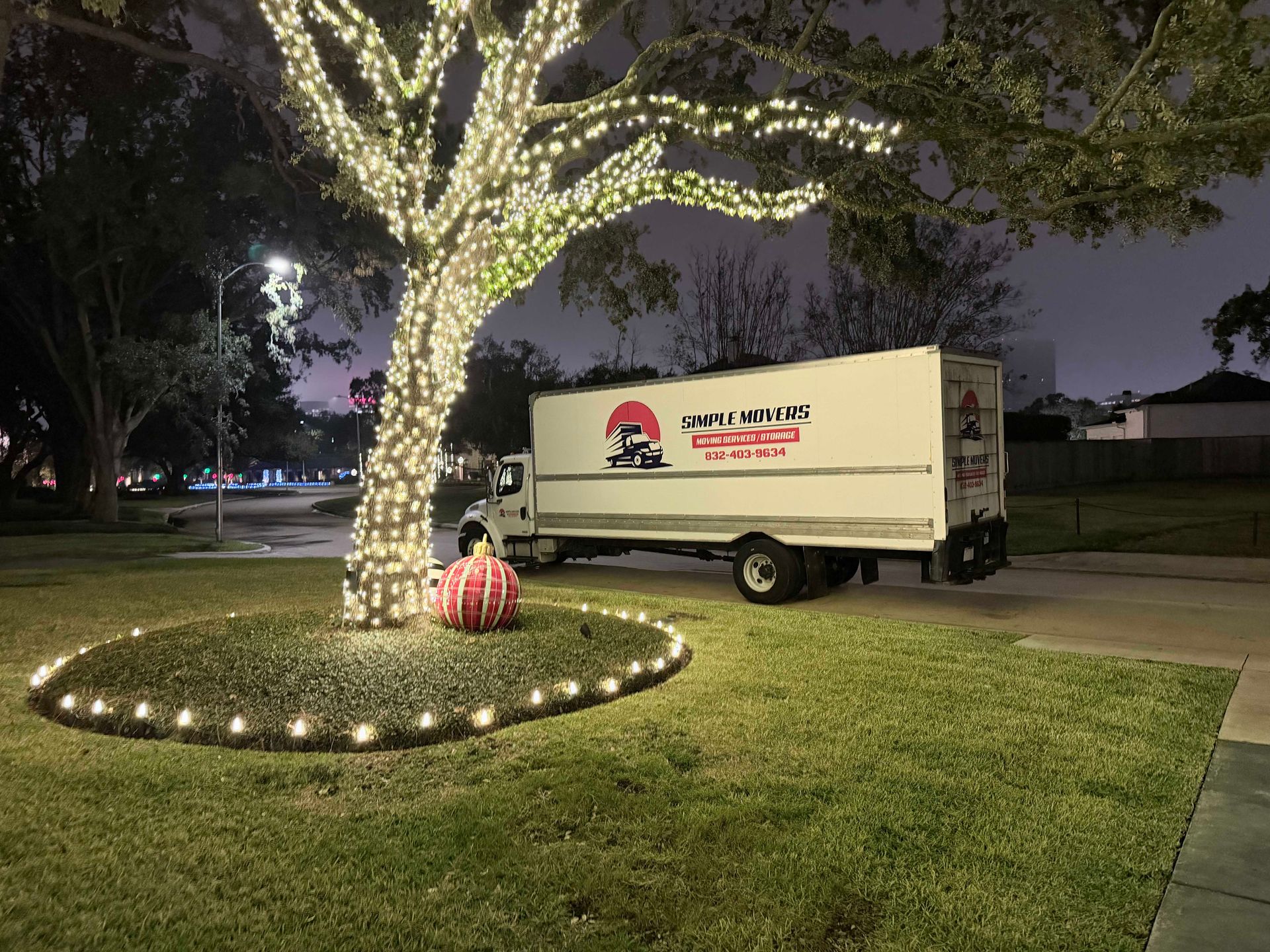 White moving truck parked under a tree with Christmas lights at night.