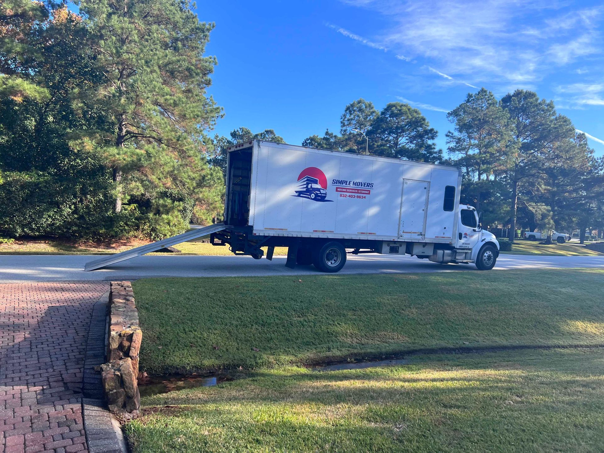 A white moving truck parked on a residential street with a ramp down. The logo is visible on the side.