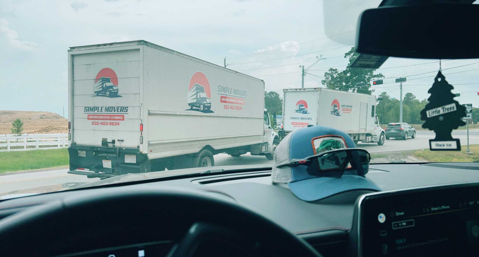 View from a car windshield of two white moving trucks driving down a road. A hat and sunglasses sit on the dashboard.