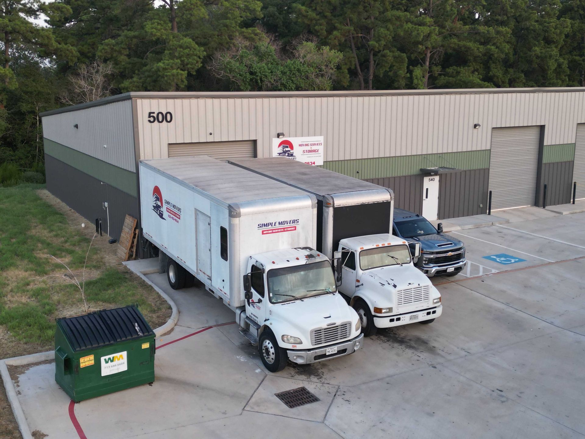 Trucks parked at a commercial building with loading dock. Green dumpster, and tree line visible.