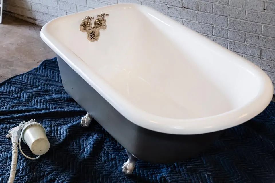 Clawfoot bathtub, white interior and gray exterior, on a blue tarp, near a paint sprayer.