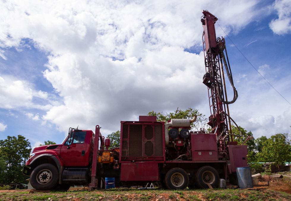 Red drilling truck with raised rig in a grassy field under a cloudy blue sky