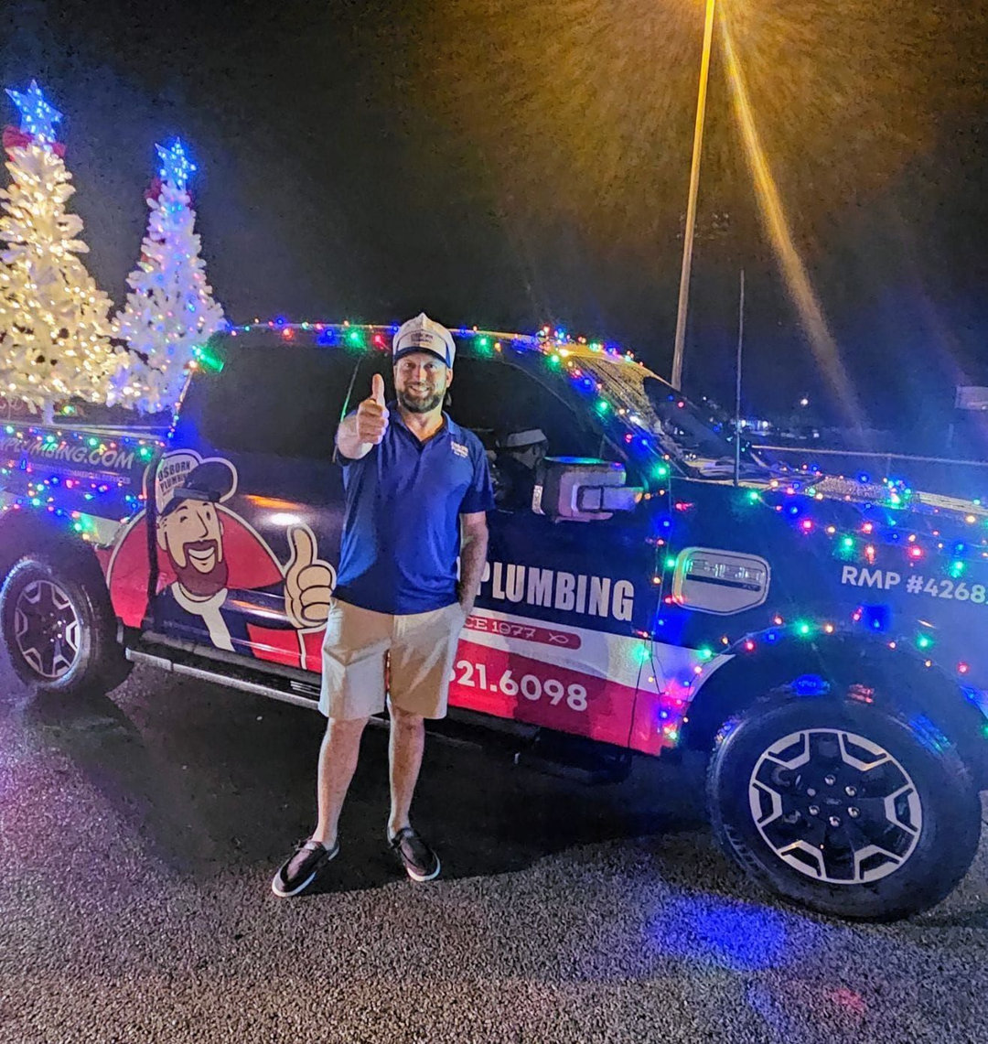 Man giving thumbs up next to a truck decorated with Christmas lights and a plumbing company logo.