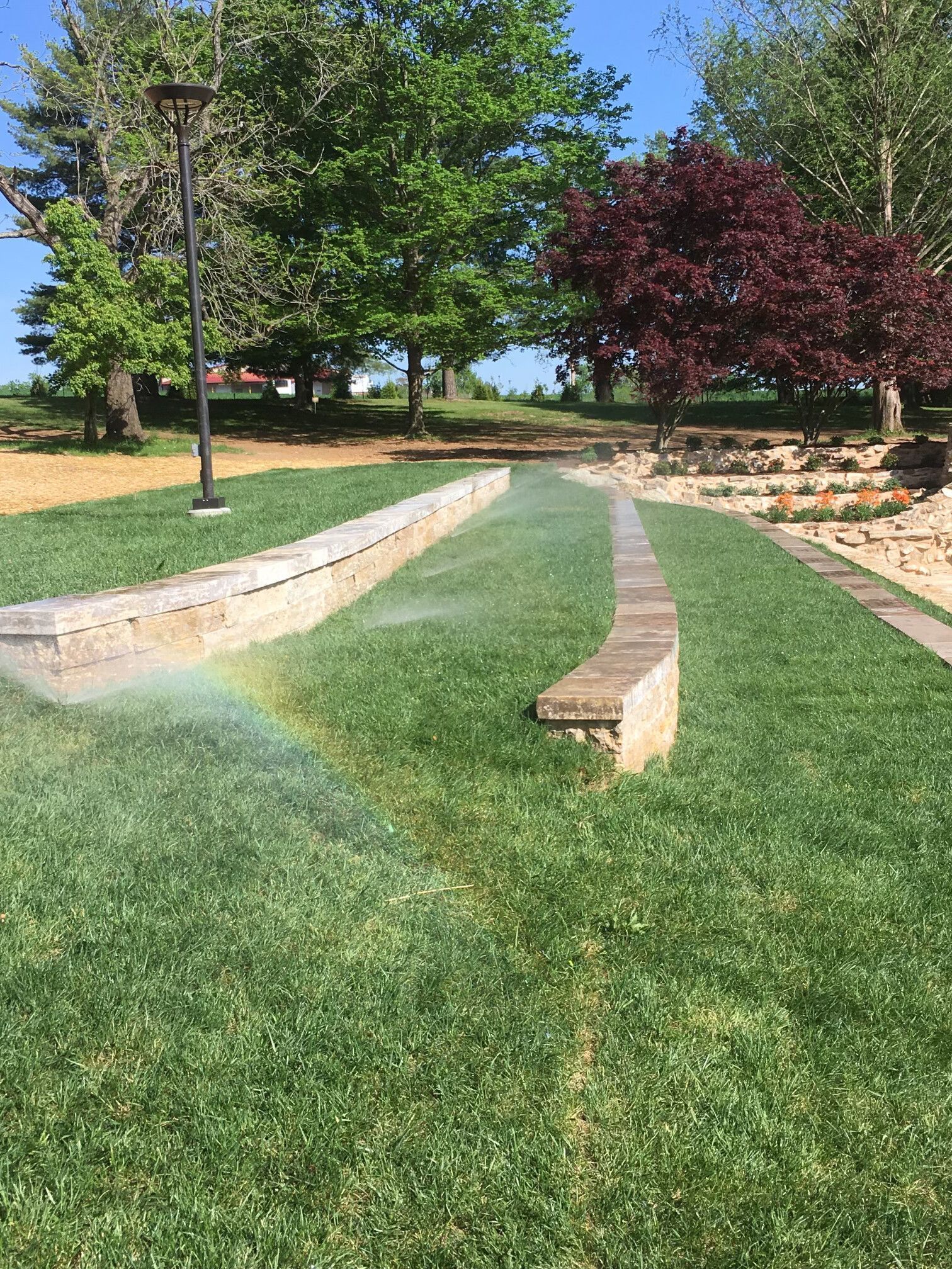 A rainbow arches over a green lawn being watered. Stone borders curve through the grass, with trees in the background.