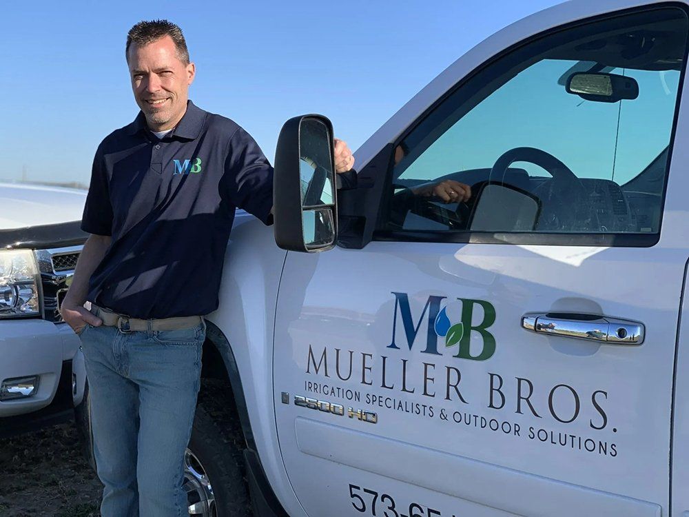 Man in blue shirt leans on white truck with "Mueller Bros." logo.