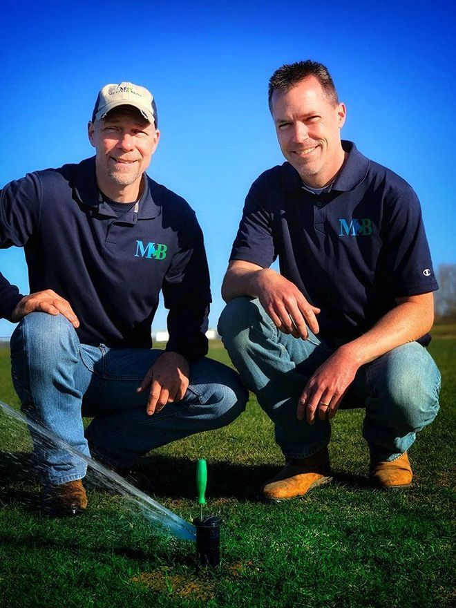 Two men in blue shirts and jeans kneel on grass near a sprinkler, smiling at the camera.