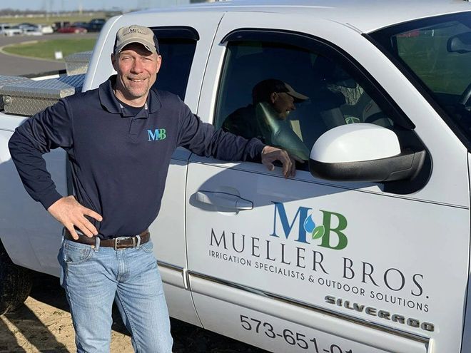 Man in blue shirt and jeans with truck; "Mueller Bros." logo on truck.