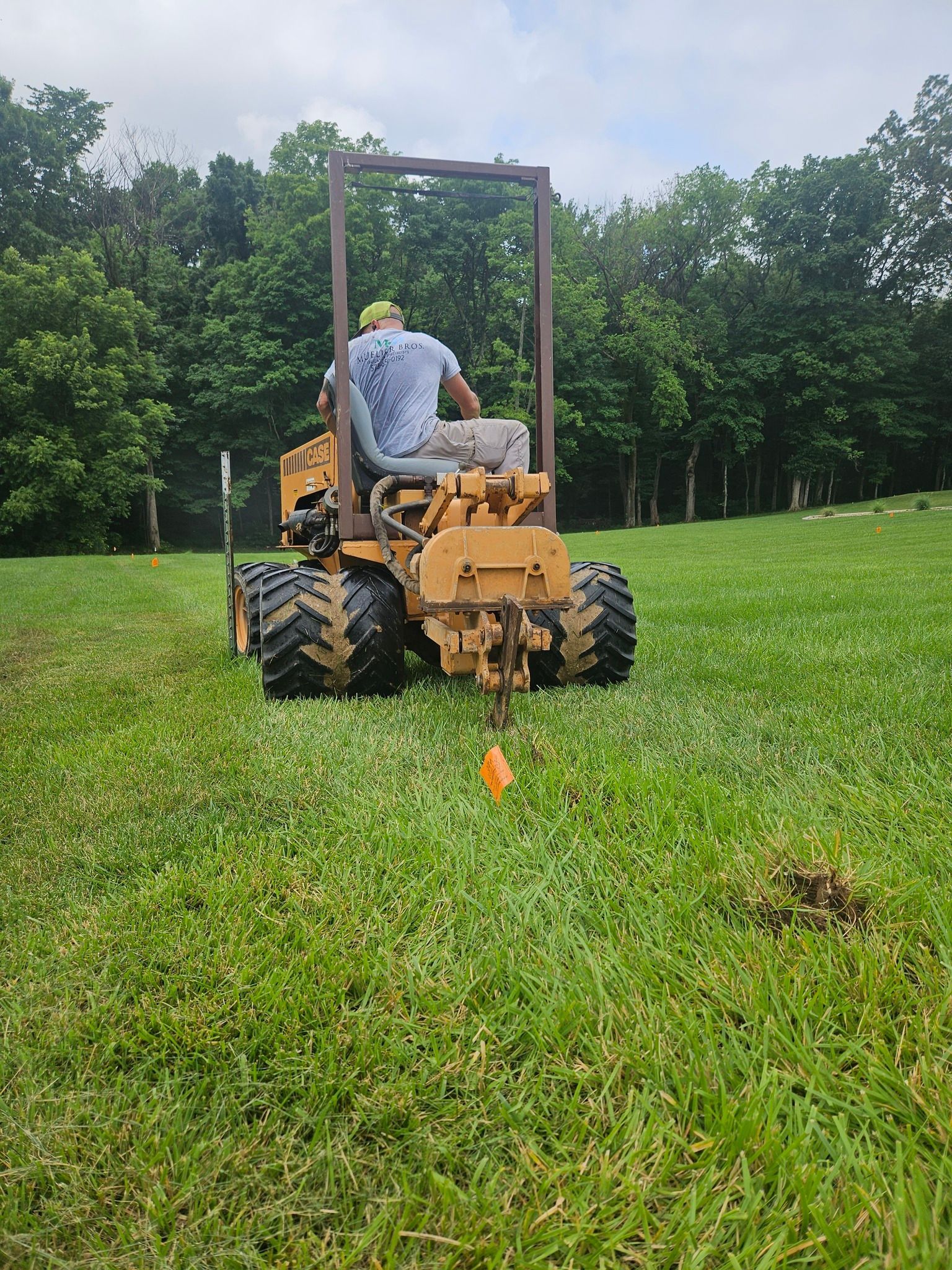 Man operating a yellow tractor in a grassy field with trees.