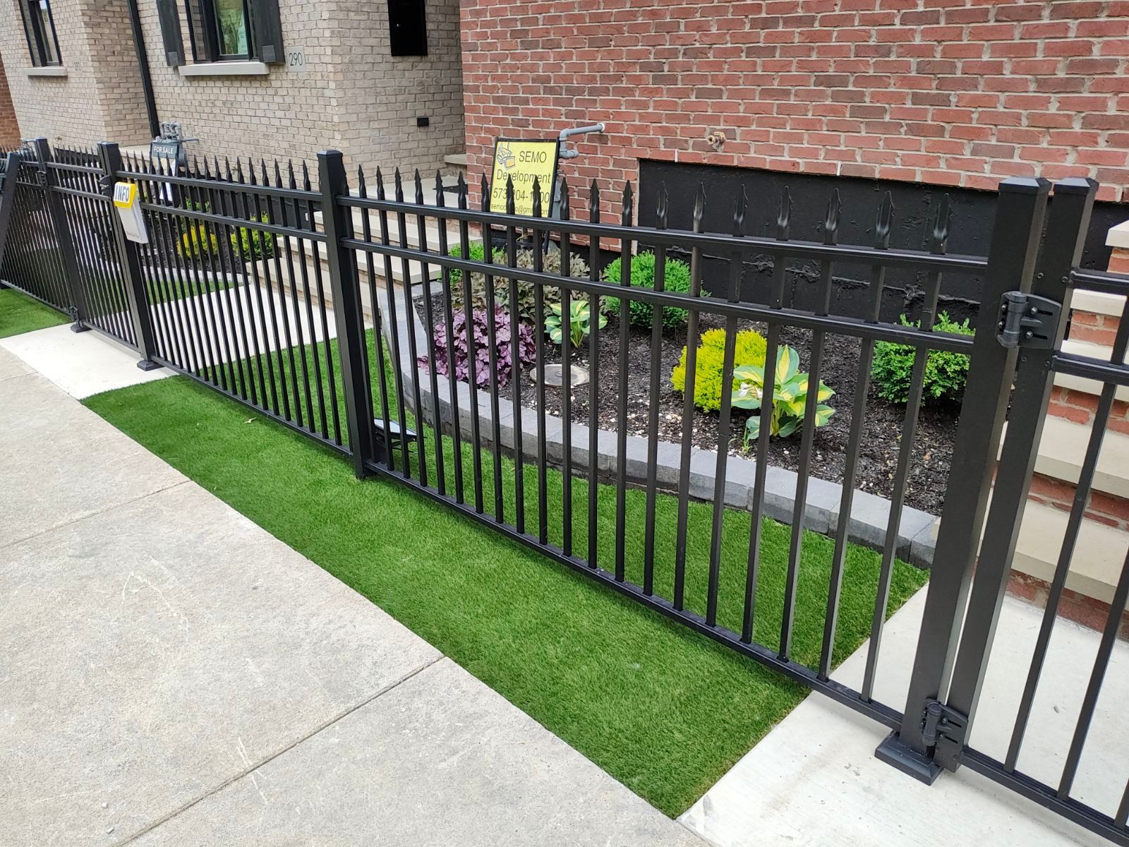 Black metal fence surrounds a small garden with artificial turf along a sidewalk, in front of a brick building.
