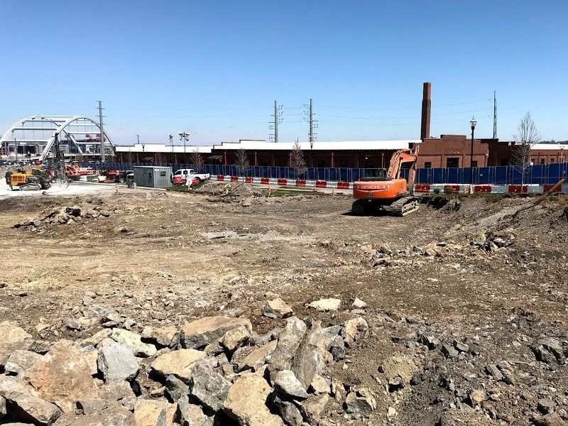 Construction site with an excavator and dirt piles, buildings and train tracks in the background.