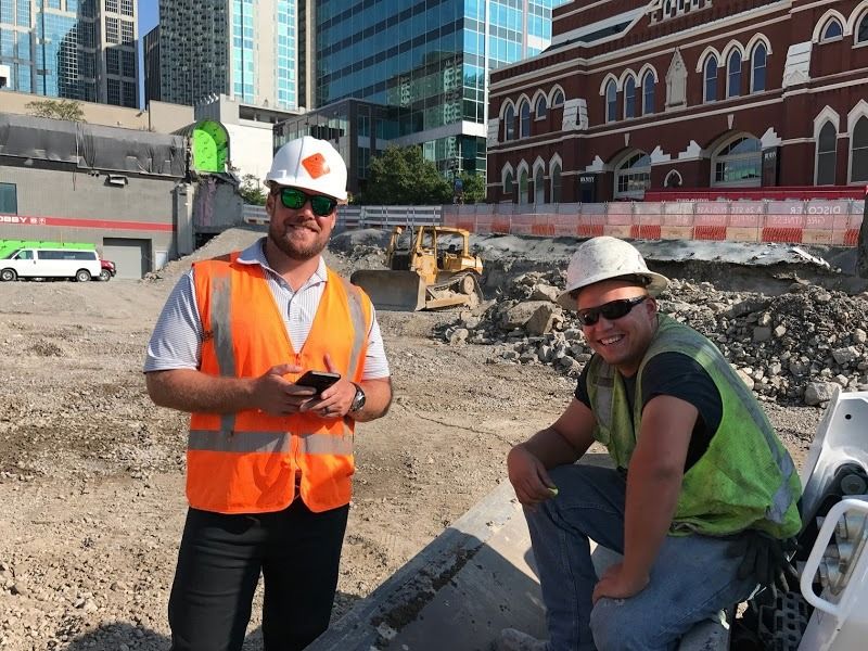Two construction workers wearing safety vests and hard hats at a construction site in front of a building.