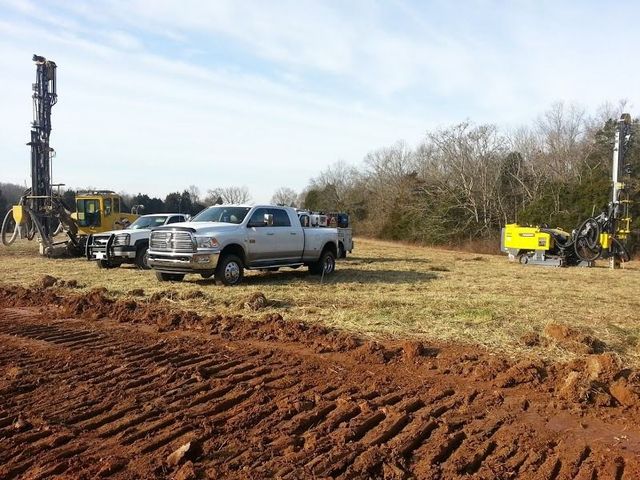 Drilling equipment and trucks in a field with a cloudy sky. Earthmoving equipment on the foreground.
