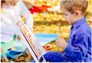 Two children painting on small canvases on easels while sitting outdoors among autumn leaves.