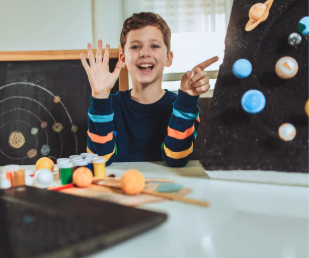 A child smiling and pointing to a DIY solar system model on a dark board, with planetary crafts on the desk.