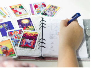 A hand holds a blue pen over an open planner, surrounded by colorful tarot cards spread out on a white surface.