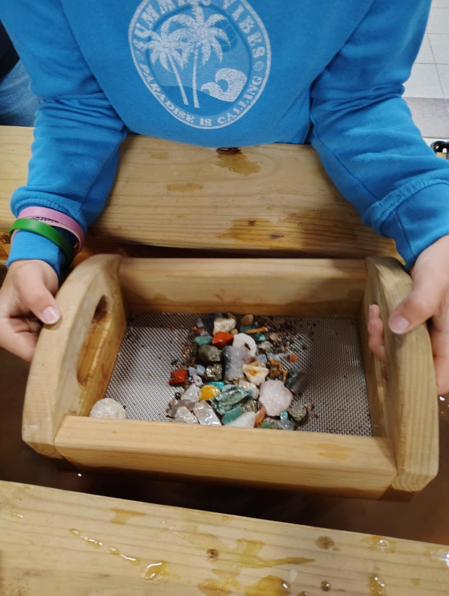 A person in a blue shirt holds a wooden sifter containing a collection of colorful polished gemstones. 