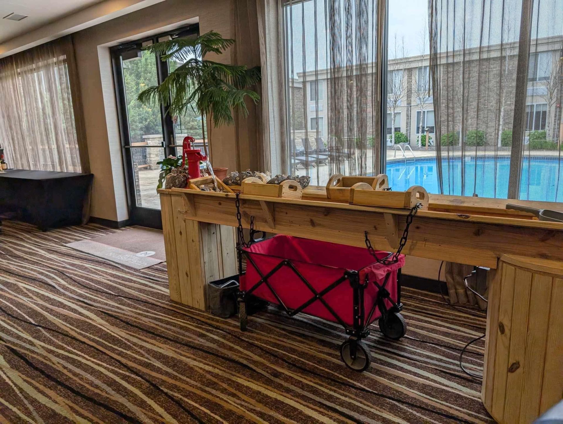 A wooden service table stands near a pool-view window, with a bright pink folding wagon stored underneath. 