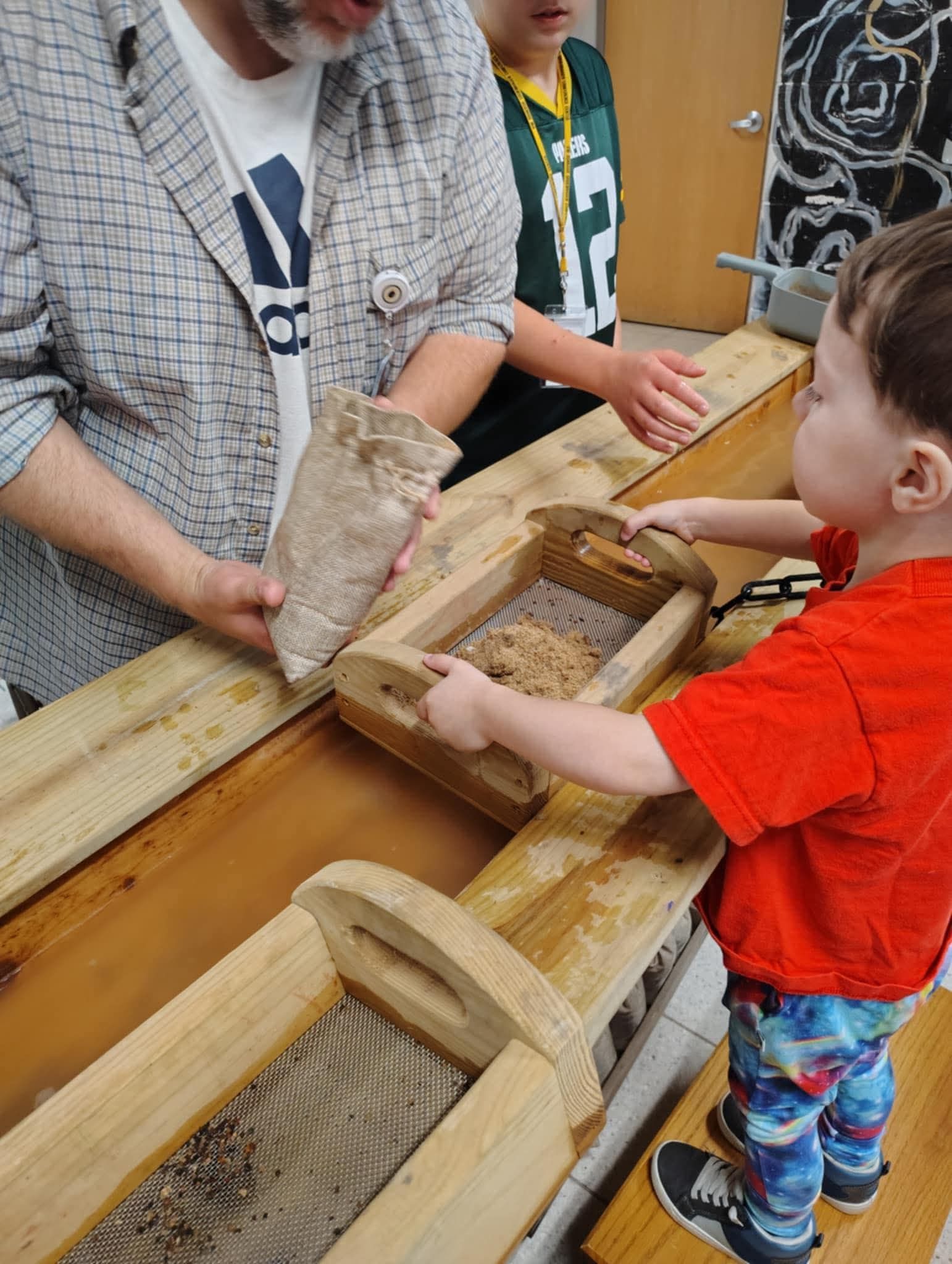 A person pours material from a small bag into a wooden sifter held by a child standing at a long wooden water trough. 